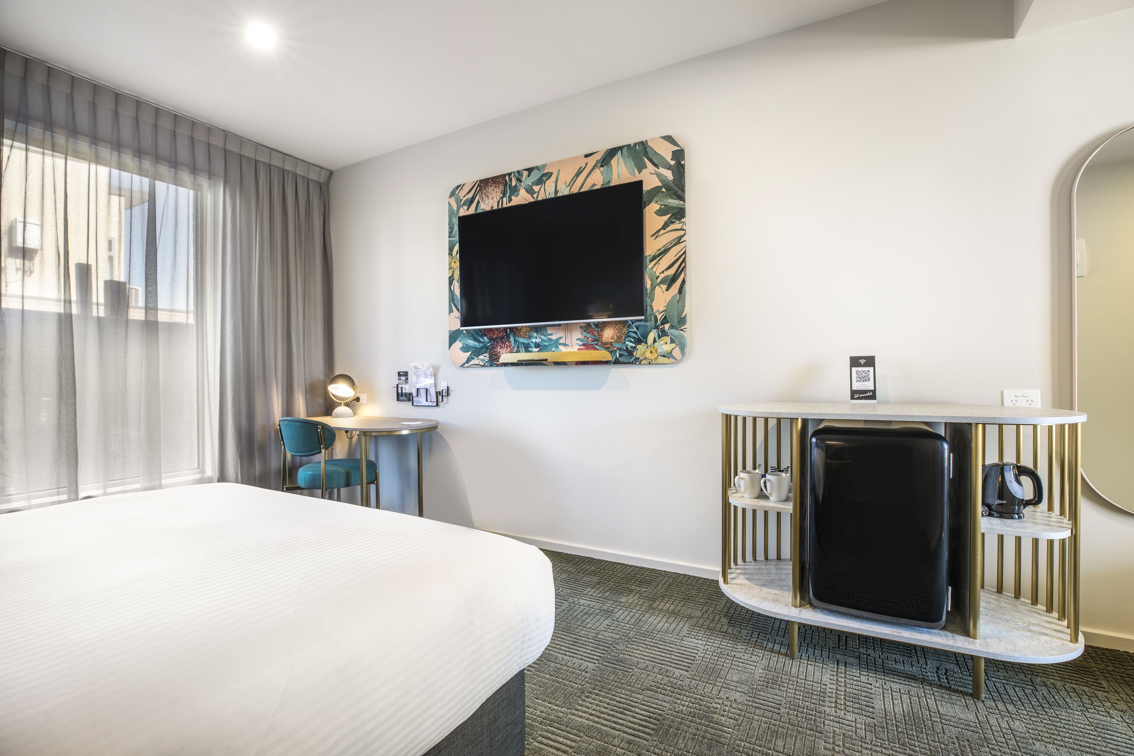 A hotel room with a bed, a desk with a lamp, a television, and a minibar.