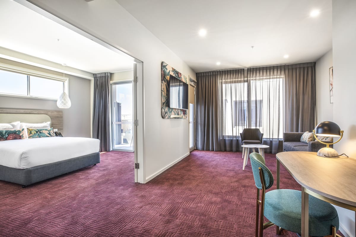 Ein Hotelzimmer mit einem großen Bett, einem Essbereich mit Tisch und Stühlen, einem an der Wand montierten Fernseher und Blick auf die Stadt durch die Fenster.
