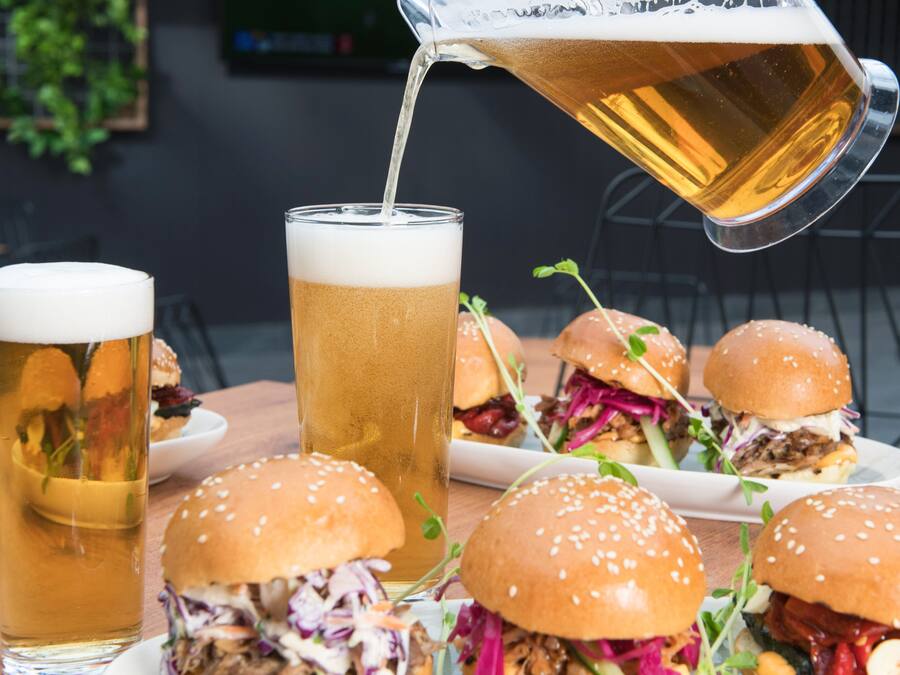 A table with a tall glass of beer, a pitcher pouring beer, and several burgers.
