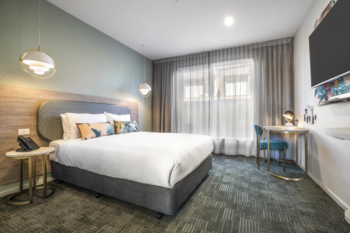 Ein Hotelzimmer mit einem großen Bett, einem Nachttisch mit Telefon, einem runden Tisch mit Lampe und einem großen Fenster mit Vorhängen.