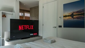 Une chambre d’hôtel avec un lit, un téléviseur affichant le logo de Netflix, un tableau représentant un coucher de soleil et une porte.