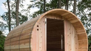 Une structure cylindrique en bois, dotée d’une porte, qui repose sur des piliers dans une zone herbeuse, avec des arbres en arrière-plan.