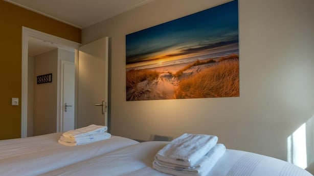 Une chambre d’hôtel avec un grand lit, un tableau représentant un coucher de soleil et une porte donnant sur une autre pièce.