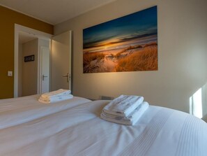 Une chambre d’hôtel avec un grand lit, un tableau représentant un coucher de soleil et une porte donnant sur une autre pièce.