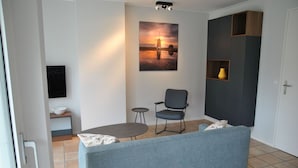 Un salon moderne avec un canapé gris, une petite table ronde, un fauteuil et un tableau représentant un coucher de soleil sur l’eau.