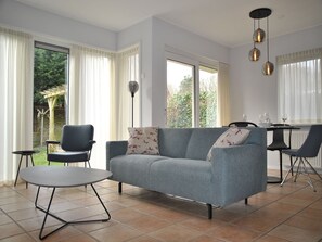 Un salon moderne avec un canapé gris, deux fauteuils noirs, une petite table ronde et une table à manger avec des chaises. De grandes fenêtres sont présentes, ainsi que des rideaux, un lampadaire et un luminaire suspendu.