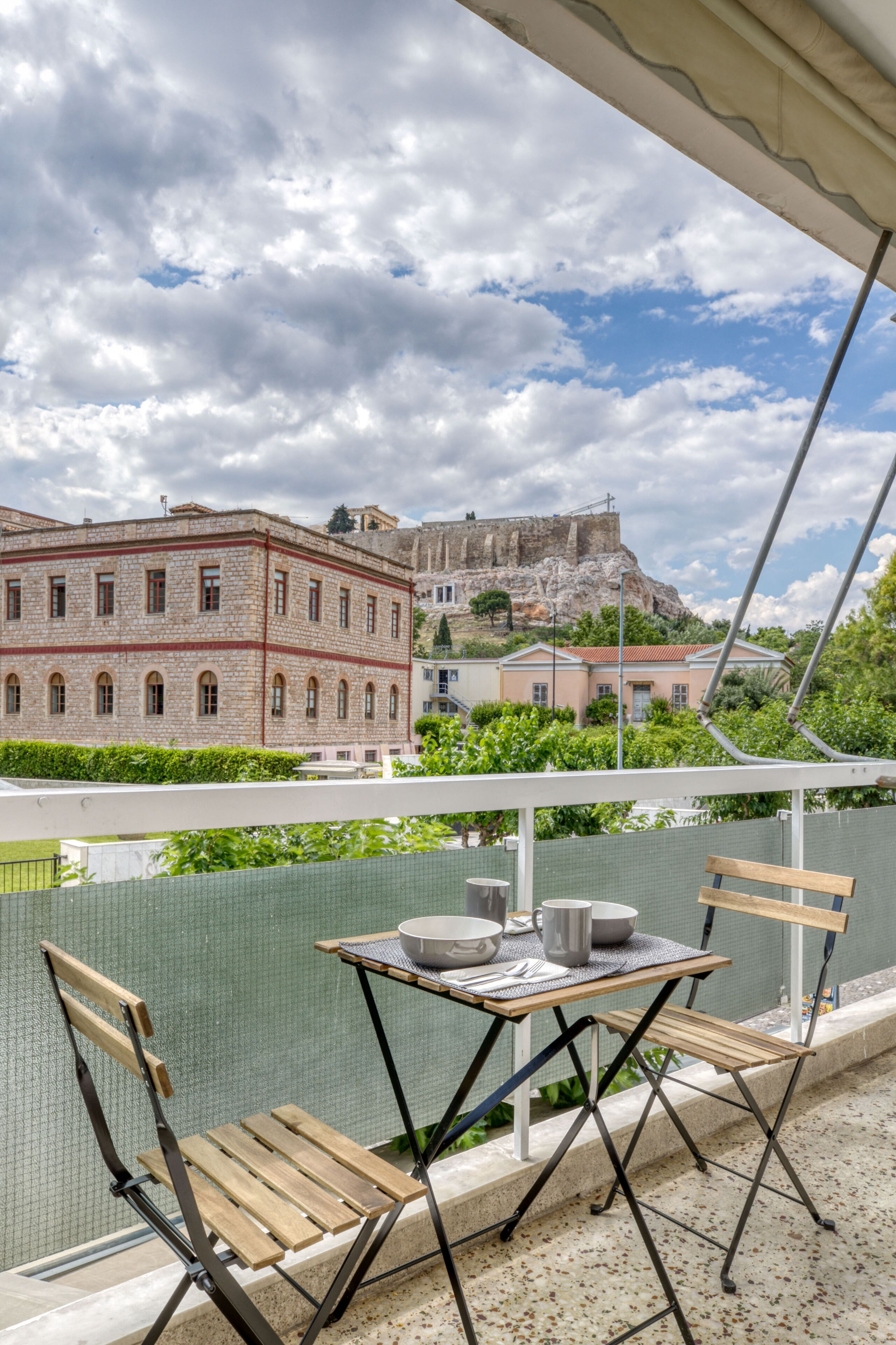 A balcony with a table and chairs overlooking a historic building and a rocky hill.