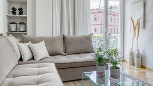En moderne stue med en beige sofa, et glasbord og udsigt til en rød bygning gennem et stort vindue.