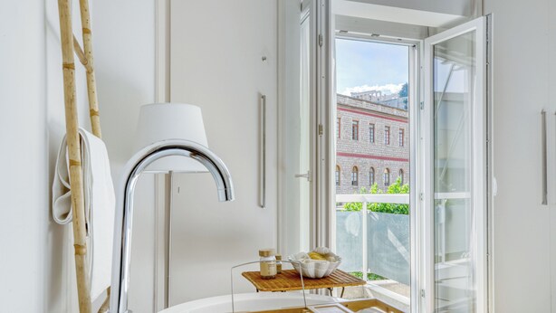 Et moderne hotelværelse med et stort vindue, et trænatbord, en hvid lampe og udsigt til en historisk bygning.