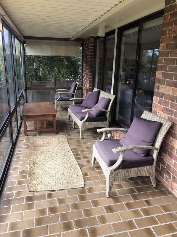 A covered patio with wicker chairs and a wooden table, a brick floor, and large windows.