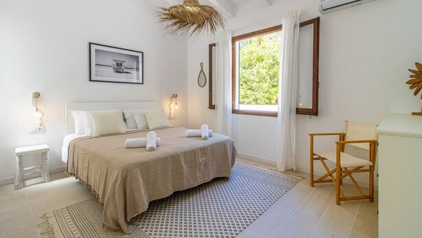 Quarto com cama, uma cadeira, mesa pequena, luminária pendente, quadro emoldurado e janela com cortinas.