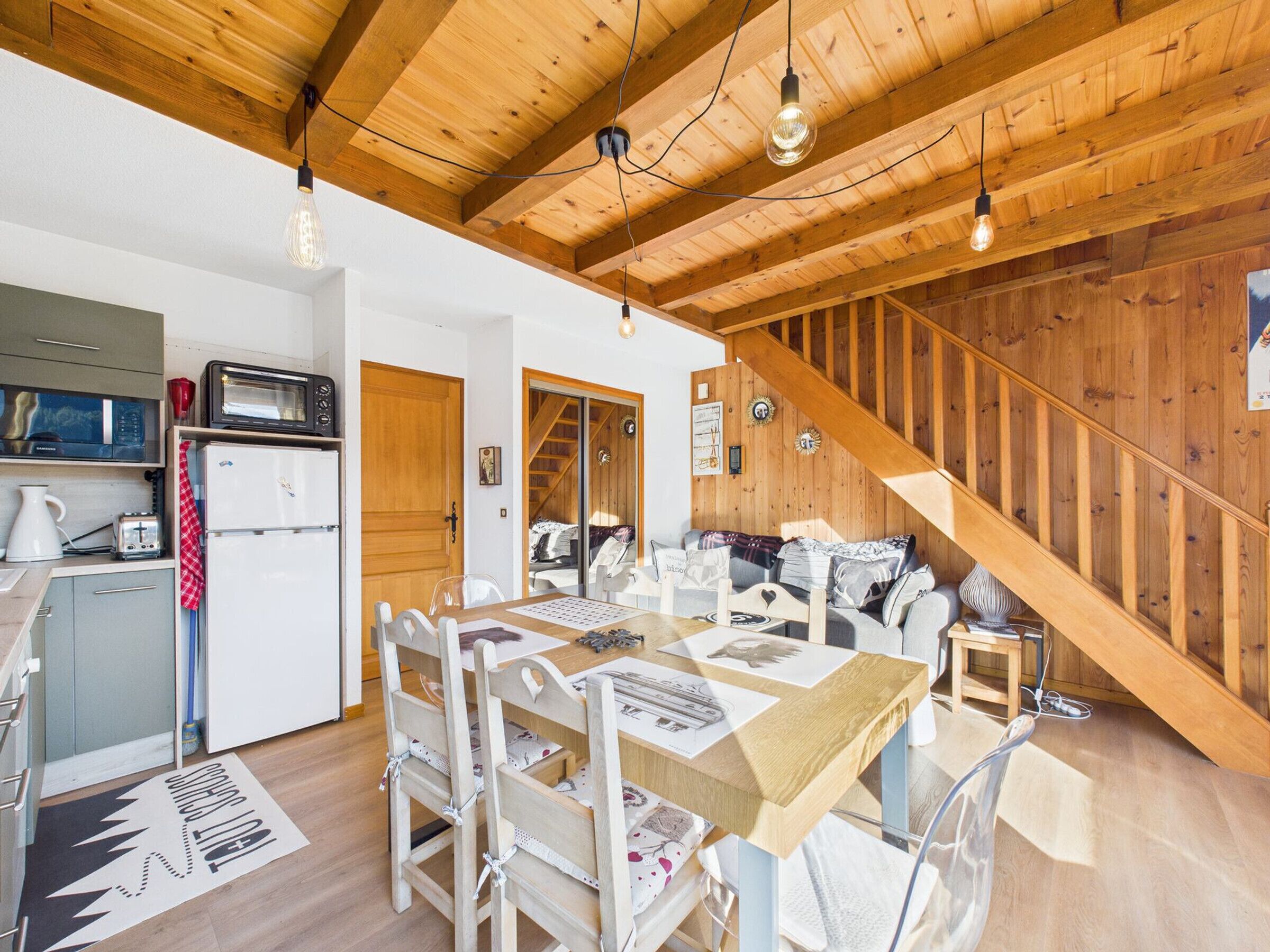 A cozy cabin interior with a wooden ceiling, a dining area with a table and chairs, a kitchen with a refrigerator and microwave, and a staircase leading to a loft bedroom.