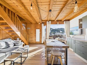 A cozy cabin interior with a wooden ceiling, a dining area, a kitchen, and a view of a snowy landscape.