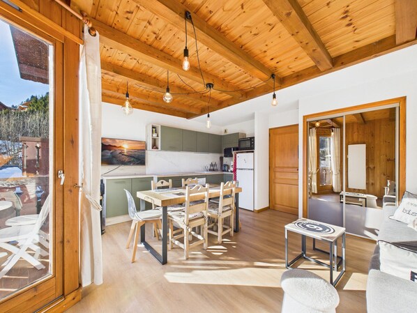 A spacious kitchen with wooden beams, a dining area with a table and chairs, and a view of the outdoors through a glass door.