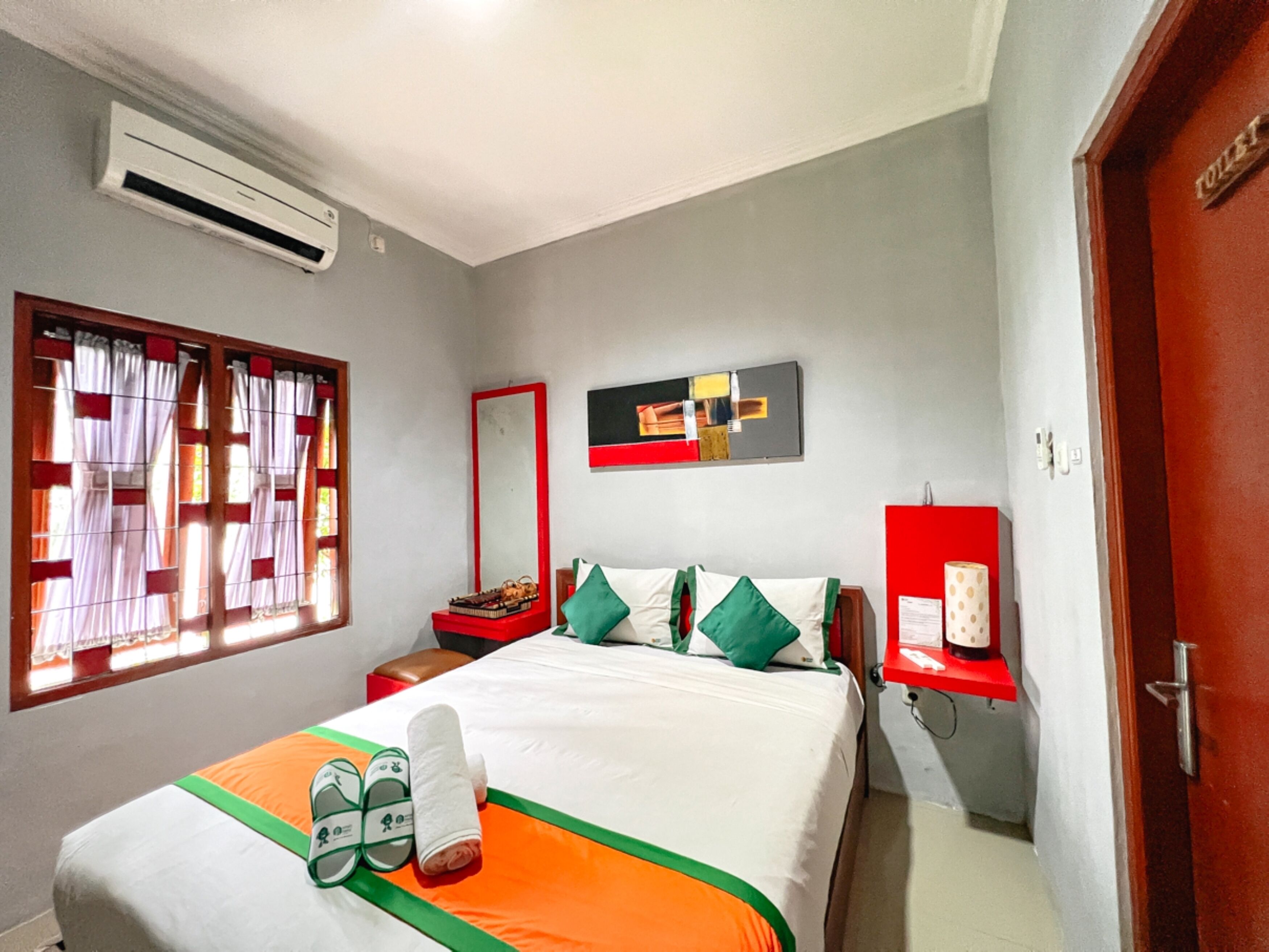 A hotel room with a bed, a nightstand, a mirror, a red shelf with a telephone, and a window with curtains.