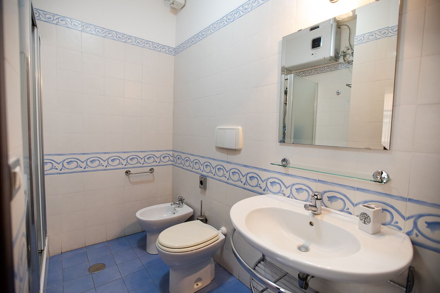 Une salle de bain comprenant des toilettes, un bidet et un lavabo, avec des carreaux de décoration bleus et une étagère en verre.