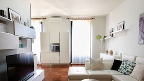 A modern living room with a flat-screen TV, a white sofa, and a kitchen area with a built-in oven and microwave. - Agatea a due passi dal Duomo by Wonderful Italy (Catania)