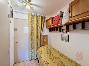 Ein Schlafzimmer mit einem Bett, einer Holzablage mit einer roten Tasche, einer Wandablage und einer Tür mit Lichtschalter.