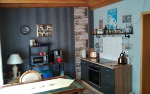 Une cuisine compacte équipée d’un four à micro-ondes, d’un grille-pain, d’une bouilloire et d’une machine à café. On y trouve une petite table à manger avec une chaise, ainsi qu’une étagère murale où sont rangés divers objets.