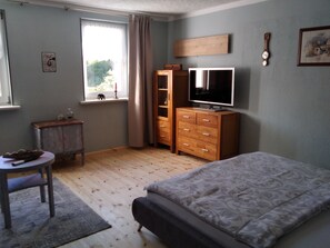 Une chambre à coucher avec un lit, une télévision, une armoire en bois, une petite table avec un bol, et une fenêtre avec des rideaux.