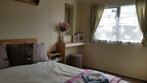 A neatly made bed with a pink and white bedspread, a small bedside table with a vase of flowers, and a window with patterned curtains. - Pool-garden-2br-parking-wifi-sleeps6 (Paignton)