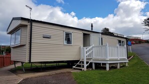 A mobile home with a deck and a chimney, located in a grassy area with a clear sky. - Pool-garden-2br-parking-wifi-sleeps6 (Paignton)