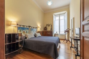 A bedroom with a bed, wooden floor, a desk, a chair, a lamp, a window, and a painting on the wall.
