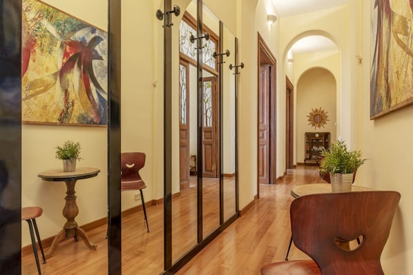A hallway with wooden flooring, a round table with a potted plant, a modern chair, and a large abstract painting on the wall. - GuestHost - Elegant 110 sqm apartment, which can accommodate up to 3 people, located on the fourth floor of a period building with lift (not suitable for the disabled). The property, in the Traffic Restricted Area (ZTL), is located in the heart of the hi (Roma)