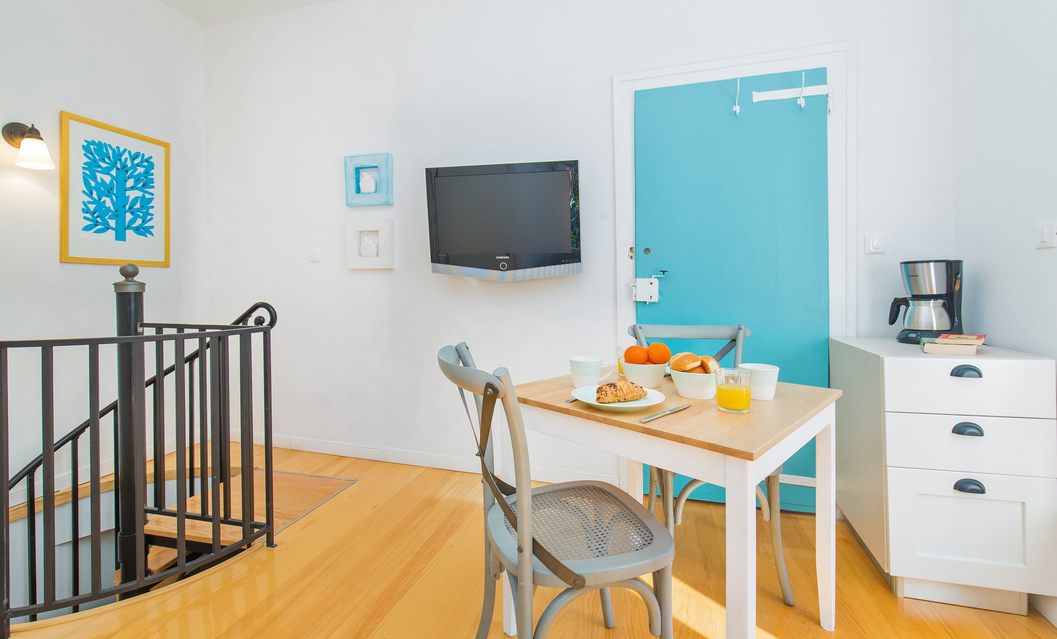 Una stanza piccola e luminosa con pavimento in legno, un tavolo da pranzo con sedie, una televisione, una macchina da caffè e una porta con cornice blu.
