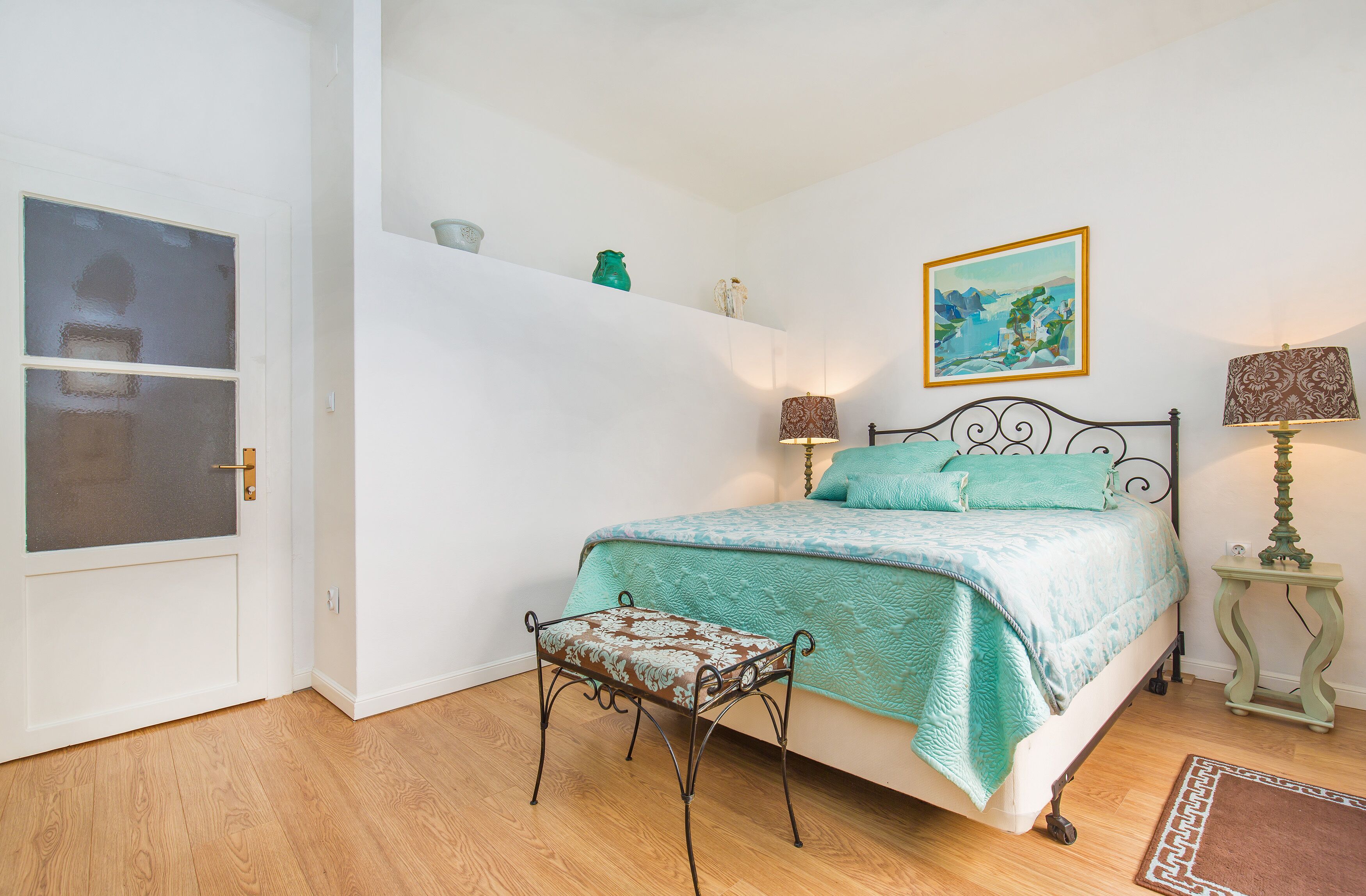 A bedroom with a bed, a bench, a door, and a shelf with decorative items.