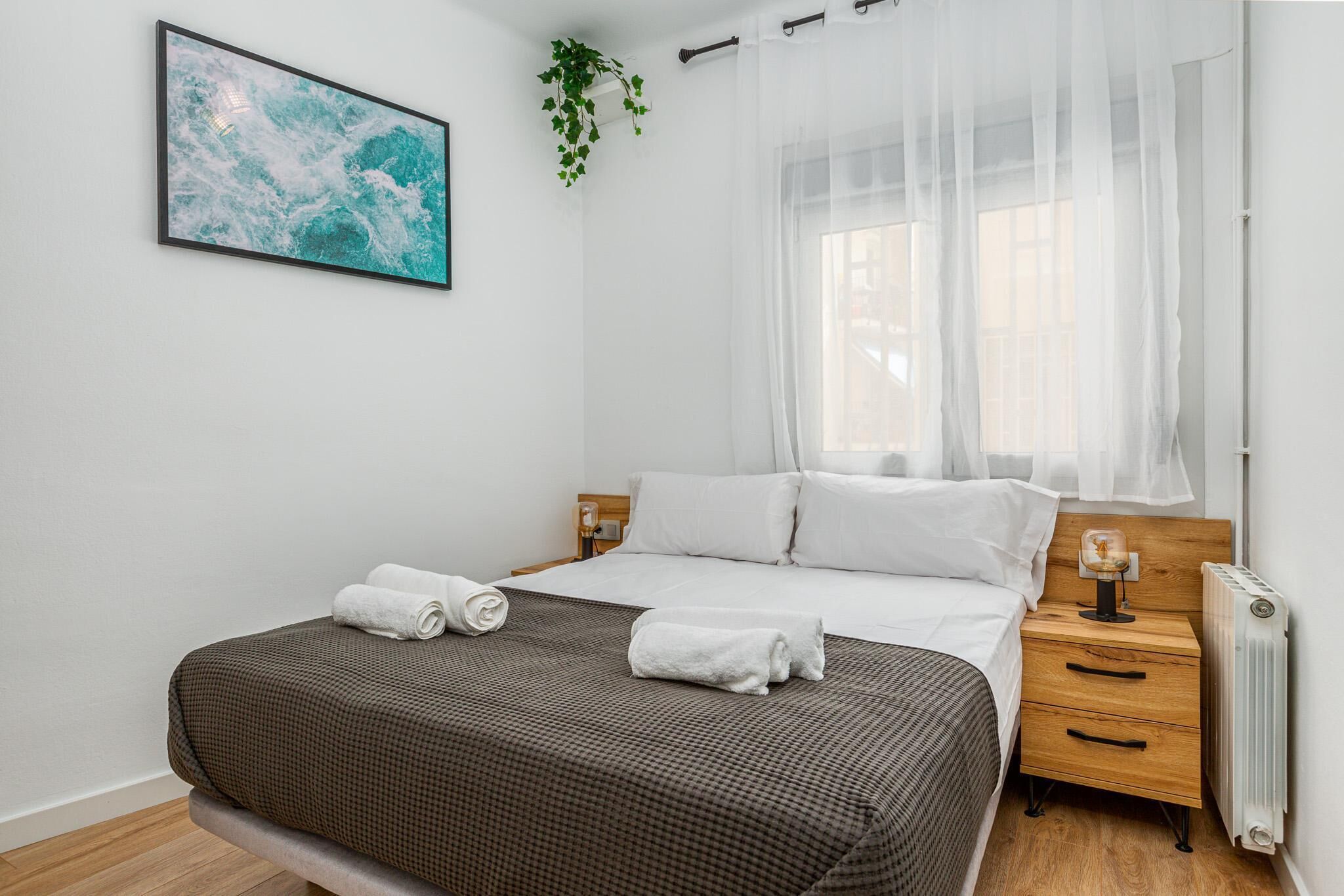 Quarto com cama, mesa de cabeceira, radiador, janela com cortinas leves e uma planta pendurada na parede, além de um quadro.