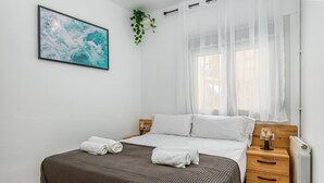 Une chambre à coucher comprenant un lit, une table de chevet, une commode, un radiateur, une fenêtre avec des rideaux légers, une plante fixée au mur et un tableau encadré.
