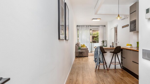 Une cuisine moderne avec un plancher en bois, un coin repas avec deux tabourets de bar, et une vue sur un salon doté d’un canapé et de rideaux.