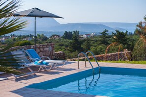 A swimming pool with lounge chairs and an umbrella, surrounded by stone walls and greenery, with a view of distant mountains. - Villa Rafaella (Apokoronas)
