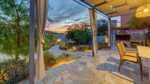 An outdoor patio with stone flooring, a dining table, chairs, and a grill. - Poseidonia Syros Cozy House (Poseidonia)
