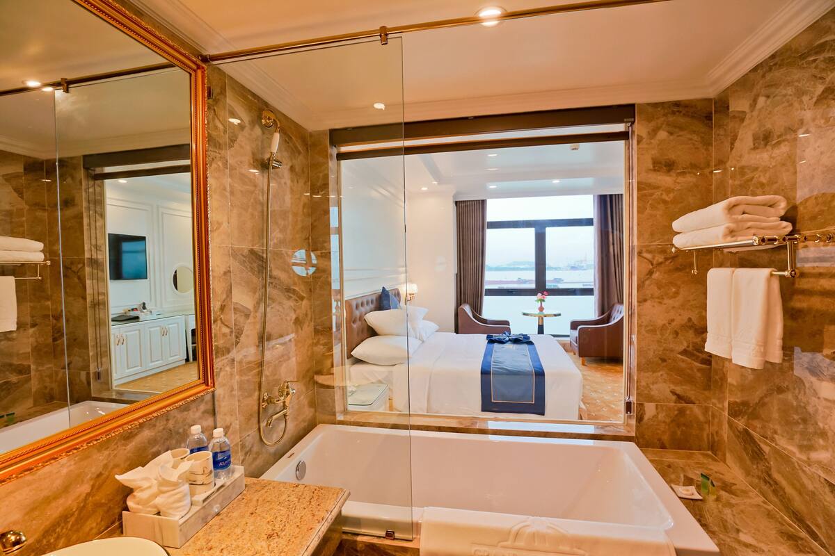 A modern hotel bathroom with a large mirror, a bathtub, a shower, and a view of a bedroom with a bed and a window.
