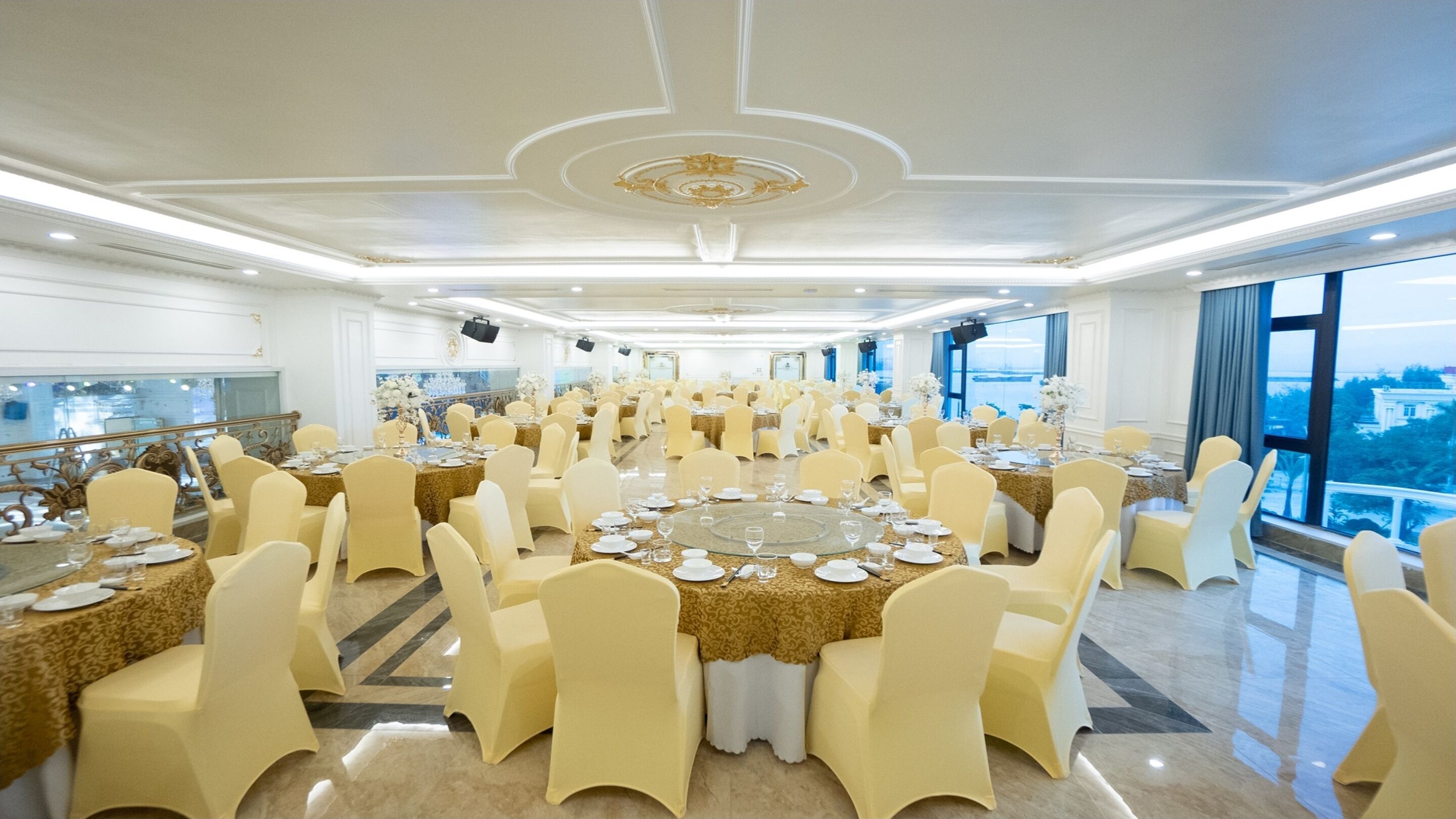 A banquet hall with round tables, chairs, and a view of the city through large windows.