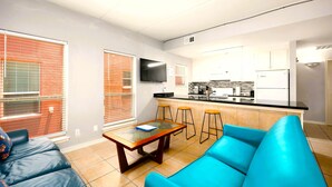 A modern living room with a blue sofa, a wooden coffee table, and a kitchen area with a black countertop and white cabinets. - Rayo del Sol II - 201 - WiFi, Walk to the beach (South Padre Island)