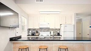A modern kitchen with white cabinets, a black countertop, and three wooden stools. - Rayo del Sol II - 201 - WiFi, Walk to the beach (South Padre Island)