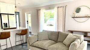 Una sala de estar moderna con un sofá color beige, dos taburetes de bar, una zona de cocina y una mesa de comedor.