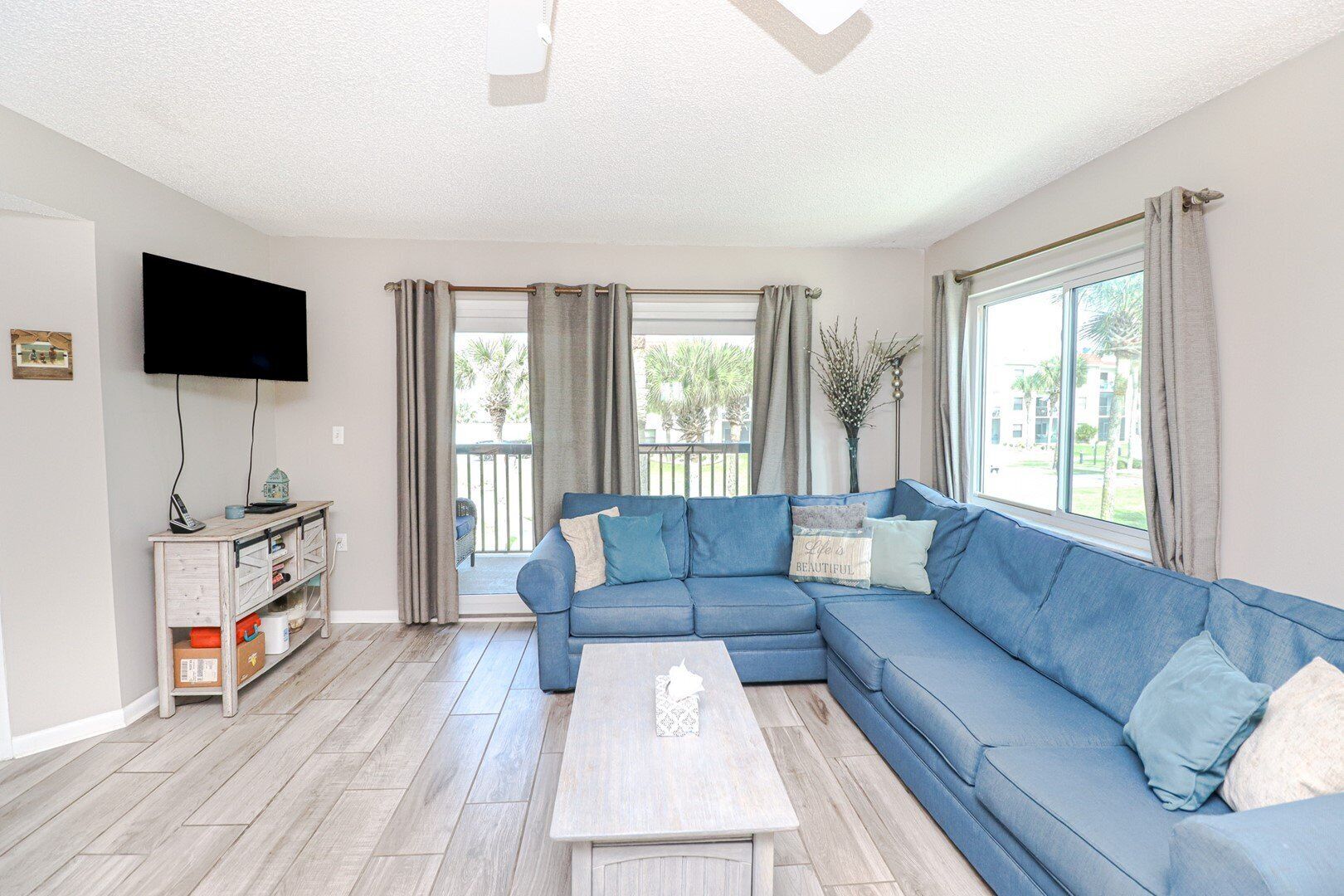 A living room with a blue sectional sofa, a coffee table, a TV on the wall, and a window with curtains.