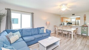 Ein Wohnzimmer mit einem blauen Sofa, einem Couchtisch und einem Essbereich mit Tisch und Stühlen. Im Hintergrund ist eine Küche mit Schränken und Geräten zu sehen.