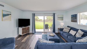 Ein Wohnzimmer mit Fernseher, blauen Sofas, einem Couchtisch und Blick auf eine Rasenfläche durch eine Glastür.