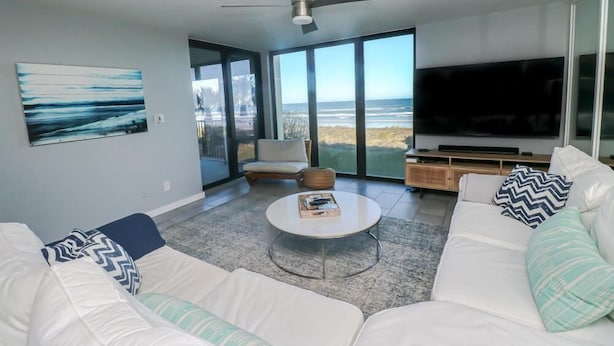 A modern living room with a large TV, white sofas, a coffee table, and a view of the ocean through large windows.