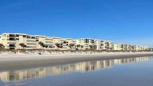 A beachfront hotel with multiple-story buildings, a sandy beach, and a calm water body reflecting the structures.