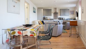 A modern kitchen with grey cabinets, a microwave, and a stove. There is a dining area with a table and chairs, and a sofa in the background. - The Anchor Apartments (Downpatrick)