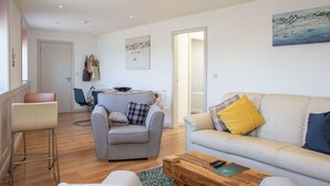 A modern living room with a grey armchair, a beige sofa, and a wooden coffee table. There is a painting on the wall and a door leading to another room. - The Anchor Apartments (Downpatrick)