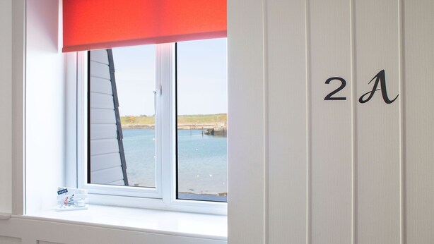 A room with a window overlooking a body of water, a door with the number 21, and a small table with a 'BEACH' sign. - The Anchor Apartments (Downpatrick)