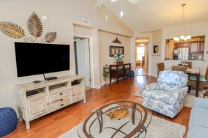 Amplia sala de estar con televisor de pantalla plana, mueble de entretenimiento de madera, sillón estampado y comedor con candelabro.