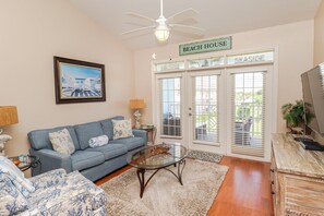 Ein Wohnzimmer mit einem blauen Sofa, einem Glastisch, einem Deckenventilator und einem Schild mit der Aufschrift 'BEACH HOUSE'.
